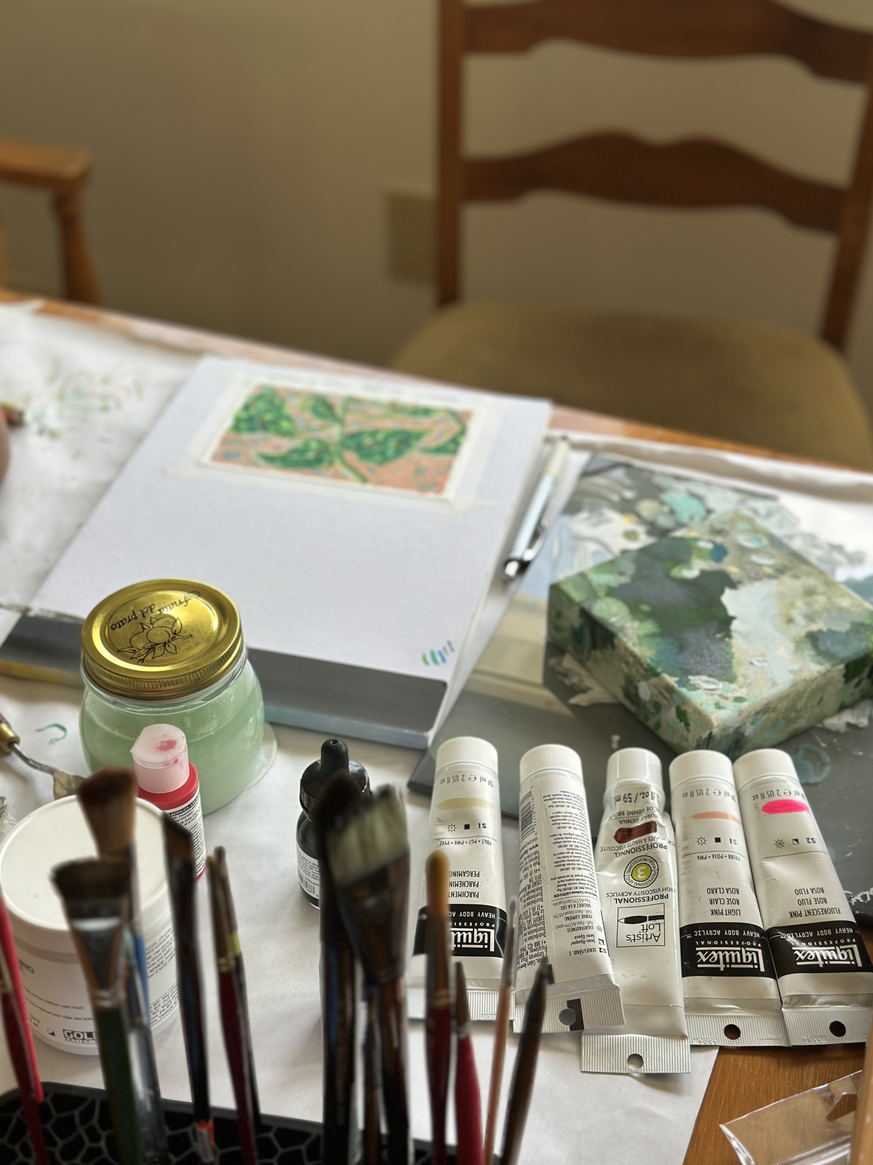 Art supplies including paint tubes, brushes, and a jar on a table with a chair in the background.